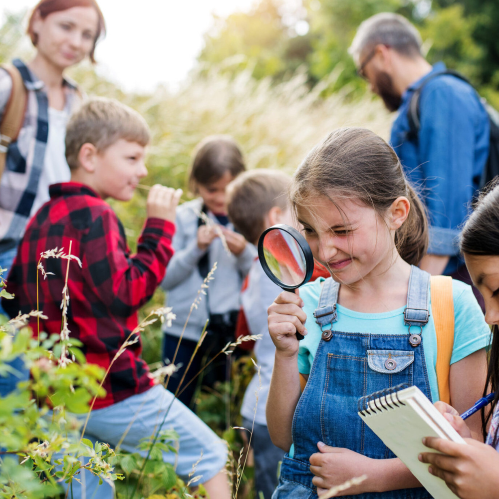 Group of adults and children hiking. Girl in front is using a magnifying glass to look at nature.